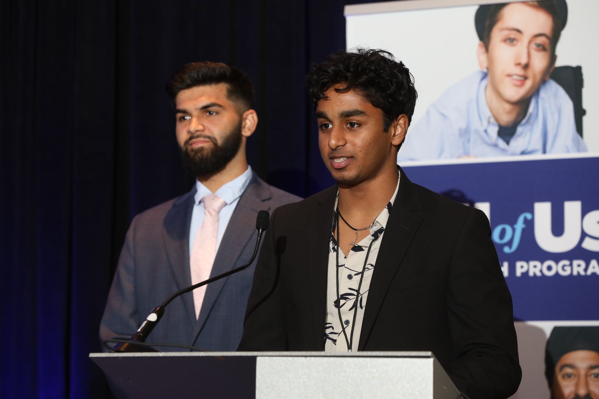 Photo from the CPGI 2023 presentation of NIH All of Us research findings, two presenters speaking at a podium with an All of Us Research Program banner in the background.