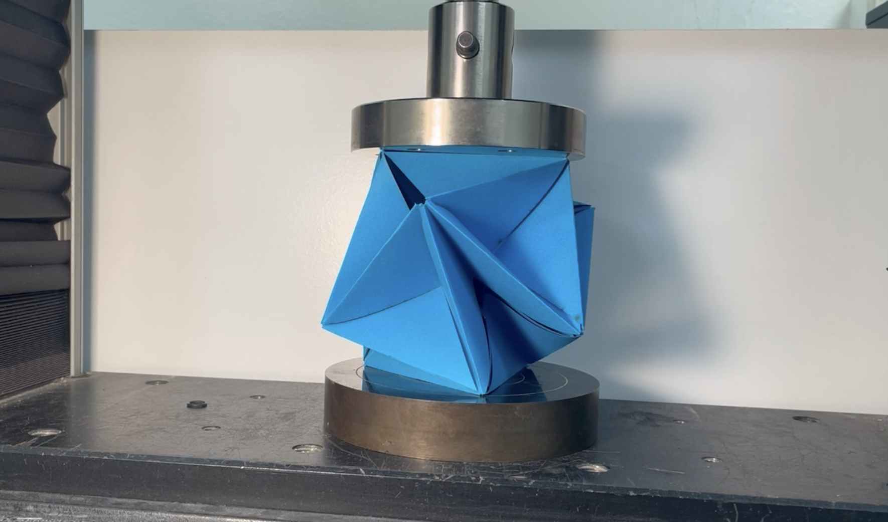 Photo of a folded, faceted geometric structure made of blue paper mounted in a mechanical testing machine between two metal compression plates.